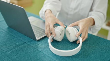 Photo for Young beautiful hispanic woman using laptop holdin headphones sitting on table at dinning room - Royalty Free Image
