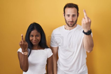 Foto de Interracial couple standing over yellow background showing middle finger, impolite and rude fuck off expression - Imagen libre de derechos