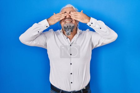 Photo for Middle age man with grey hair standing over blue background covering eyes with hands smiling cheerful and funny. blind concept. - Royalty Free Image