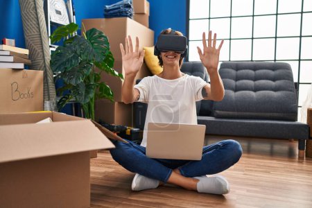 Photo for Young beautiful hispanic woman playing video game using virtual reality glasses sitting on floor at new home - Royalty Free Image
