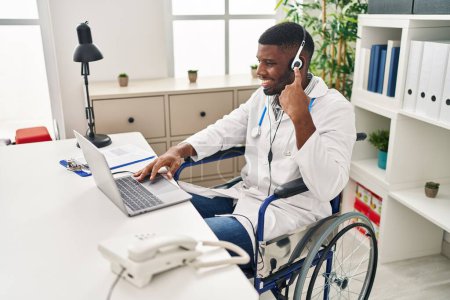 Téléchargez les photos : Afro-américain médecin homme travaillant sur rendez-vous en ligne assis sur fauteuil roulant souriant heureux pointant avec la main et le doigt - en image libre de droit