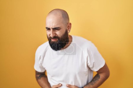 Téléchargez les photos : Jeune homme hispanique avec barbe et tatouages debout sur fond jaune avec la main sur le ventre parce que l'indigestion, la maladie douloureuse se sentent mal. concept de douleur. - en image libre de droit