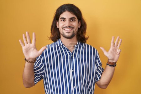 Foto de Hombre hispano con el pelo largo de pie sobre fondo amarillo mostrando y señalando hacia arriba con los dedos número nueve mientras sonríe confiado y feliz. - Imagen libre de derechos