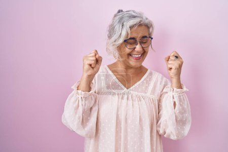 Téléchargez les photos : Femme d'âge moyen avec les cheveux gris debout sur fond rose excité pour le succès avec les bras levés et les yeux fermés célébrant la victoire en souriant. concept gagnant. - en image libre de droit