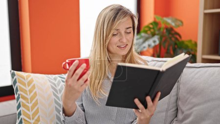 Photo for Young blonde woman reading book and drinking coffee sitting on sofa at home - Royalty Free Image
