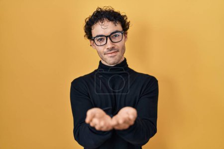 Foto de Hispanic man standing over yellow background smiling with hands palms together receiving or giving gesture. hold and protection - Imagen libre de derechos