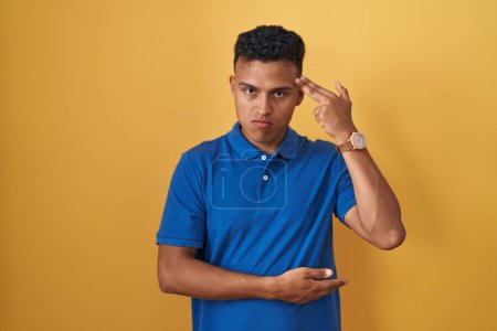 Foto de Joven hispano parado sobre fondo amarillo disparando y matándose apuntando de la mano y los dedos a la cabeza como arma, gesto suicida. - Imagen libre de derechos
