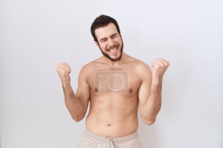 Photo for Young hispanic man standing shirtless over white background very happy and excited doing winner gesture with arms raised, smiling and screaming for success. celebration concept. - Royalty Free Image