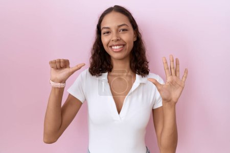 Foto de Mujer hispana joven vistiendo camiseta blanca casual mostrando y apuntando hacia arriba con los dedos número seis mientras sonríe confiada y feliz. - Imagen libre de derechos