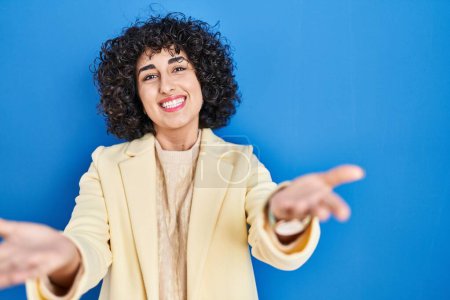 Téléchargez les photos : Jeune femme brune avec des cheveux bouclés debout sur fond bleu souriant joyeuse offrant des mains donnant assistance et acceptation. - en image libre de droit
