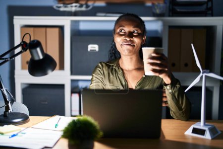Foto de Mujer africana trabajando usando computadora portátil por la noche sonriendo mirando hacia un lado y mirando hacia otro pensando. - Imagen libre de derechos
