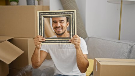 Foto de Joven hispano sosteniendo marco vintage sentado en sofá sonriendo en casa nueva - Imagen libre de derechos