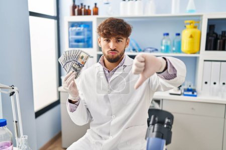 Photo for Arab man with beard working at scientist laboratory holding money with angry face, negative sign showing dislike with thumbs down, rejection concept - Royalty Free Image
