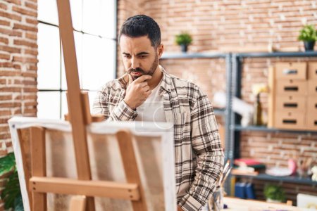 Téléchargez les photos : Jeune homme hispanique regardant dessiner avec l'expression du doute au studio d'art - en image libre de droit