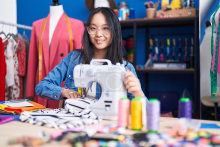 Téléchargez les photos : Jeune femme chinoise tailleur souriant confiant en utilisant une machine à coudre à l'usine de vêtements - en image libre de droit