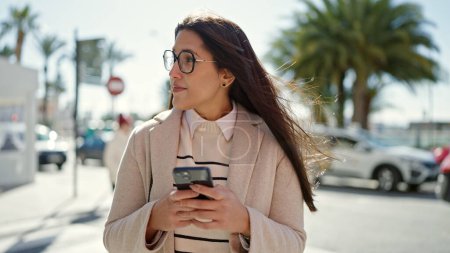 Foto de Mujer hispana hermosa joven usando teléfono inteligente con expresión seria en la calle - Imagen libre de derechos