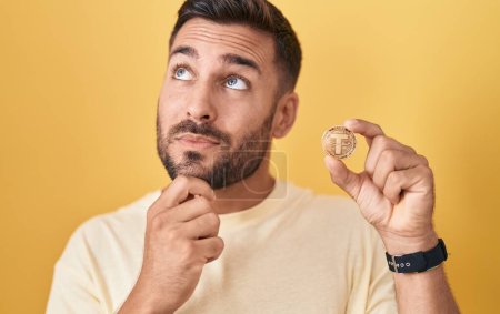 Schöner hispanischer Mann, der eine Kryptowährungsmünze in der Hand hält und ernsthaft über Fragen nachdenkt, mit der Hand am Kinn, nachdenklich über die verwirrende Idee 