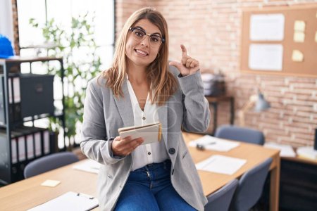 Photo for Young hispanic woman working at the office wearing glasses smiling and confident gesturing with hand doing small size sign with fingers looking and the camera. measure concept. - Royalty Free Image