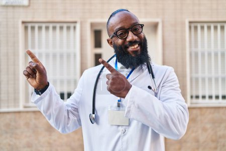 Foto de Hombre afroamericano vistiendo uniforme médico y estetoscopio sonriendo y mirando a la cámara apuntando con dos manos y dedos hacia un lado. - Imagen libre de derechos