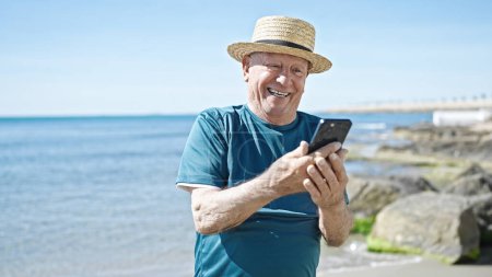 Foto de Senior hombre de pelo gris turista con sombrero de verano con teléfono inteligente en la playa - Imagen libre de derechos