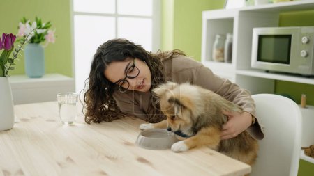 Photo for Young hispanic woman with dog sitting on table drinking water at dinning room - Royalty Free Image