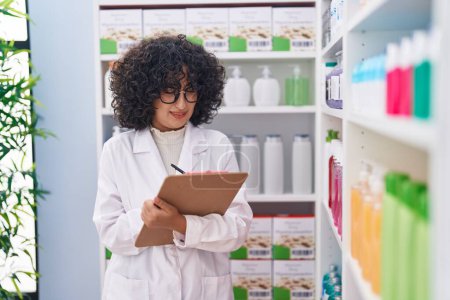 Téléchargez les photos : Jeune pharmacienne du Moyen-Orient écrivant sur un document à la pharmacie - en image libre de droit