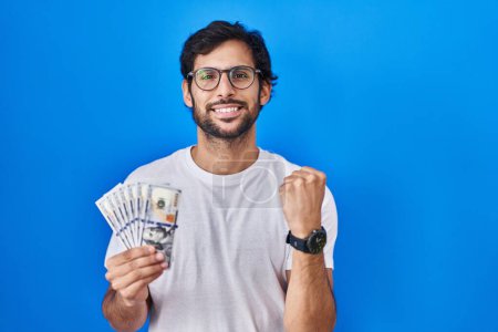 Photo for Handsome latin man holding dollars banknotes screaming proud, celebrating victory and success very excited with raised arms - Royalty Free Image