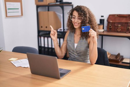 Photo for Hispanic woman with curly hair wearing call center agent headset holding credit card smiling happy pointing with hand and finger to the side - Royalty Free Image