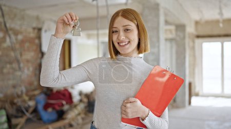 Photo for Young blonde woman real state agent holding keys of new home and clipboard at construction site - Royalty Free Image
