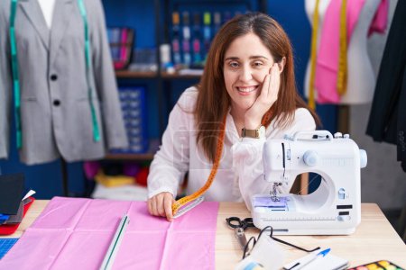 Téléchargez les photos : Jeune femme tailleur souriant confiant assis sur la table au studio de couture - en image libre de droit