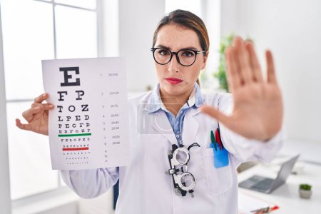 Photo for Young hispanic optician woman holding medical exam with open hand doing stop sign with serious and confident expression, defense gesture - Royalty Free Image