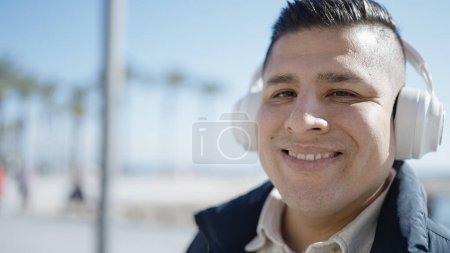Téléchargez les photos : Jeune homme hispanique écoutant de la musique au bord de la mer - en image libre de droit