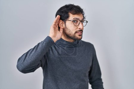 Photo for Handsome latin man standing over isolated background smiling with hand over ear listening an hearing to rumor or gossip. deafness concept. - Royalty Free Image