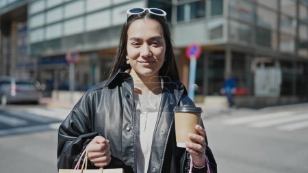 Téléchargez les photos : Jeune belle femme hispanique souriant confiant tenant sacs à provisions et café à la rue - en image libre de droit