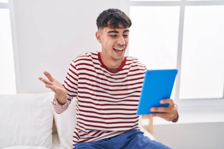 Téléchargez les photos : Jeune homme hispanique utilisant pavé tactile assis sur le lit célébrant la réalisation avec sourire heureux et expression gagnante avec la main levée - en image libre de droit