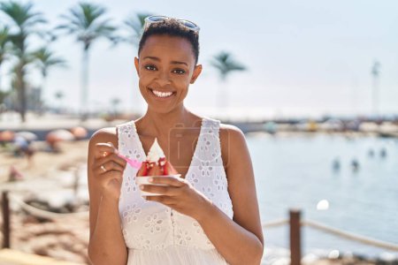 Téléchargez les photos : Femme afro-américaine souriante manger de la glace en toute confiance au bord de la mer - en image libre de droit