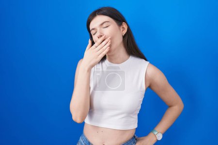 Téléchargez les photos : Jeune femme caucasienne debout sur fond bleu ennuyé bâillement fatigué couvrant la bouche avec la main. agitation et somnolence. - en image libre de droit