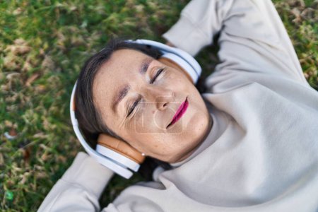 Téléchargez les photos : Femme d'âge moyen écoutant de la musique allongée sur une herbe au parc - en image libre de droit