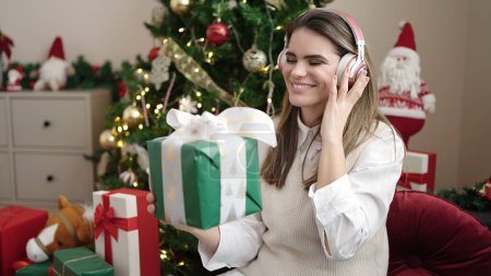 Téléchargez les photos : Jeune belle femme hispanique souriant confiant tenant cadeau de Noël portant des écouteurs à la maison - en image libre de droit