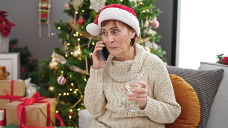 Photo for Mature hispanic woman speaking on the phone by christmas tree drinking wine at home - Royalty Free Image
