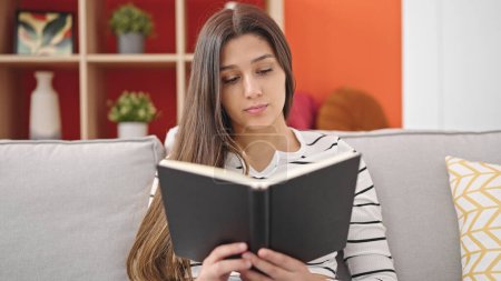 Foto de Joven mujer hispana hermosa leyendo libro sentado en el sofá en casa - Imagen libre de derechos