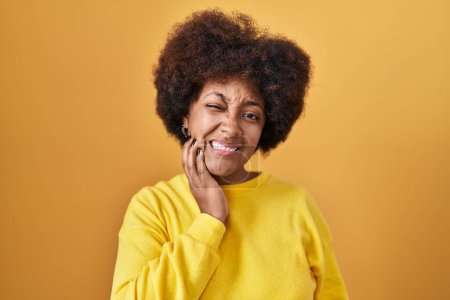 Foto de Young african american woman standing over yellow background touching mouth with hand with painful expression because of toothache or dental illness on teeth. dentist - Imagen libre de derechos