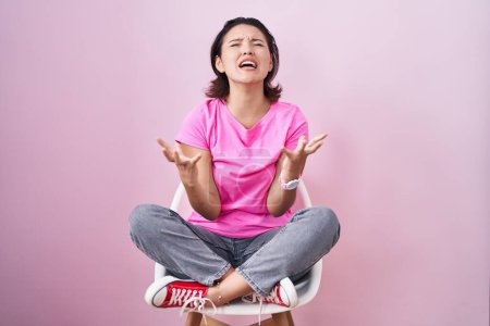 Photo for Hispanic young woman sitting on chair over pink background crazy and mad shouting and yelling with aggressive expression and arms raised. frustration concept. - Royalty Free Image
