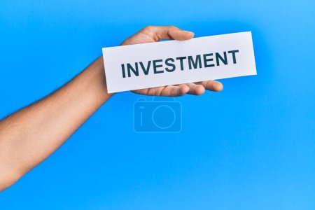 Hand of caucasian man holding paper with investment word over isolated blue background