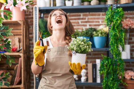 Foto de Joven mujer caucásica trabajando en floristería sosteniendo planta enojada y loca gritando frustrada y furiosa, gritando con ira mirando hacia arriba. - Imagen libre de derechos