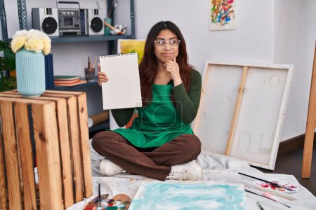 Photo for Hispanic young woman holding notebook at art studio serious face thinking about question with hand on chin, thoughtful about confusing idea - Royalty Free Image