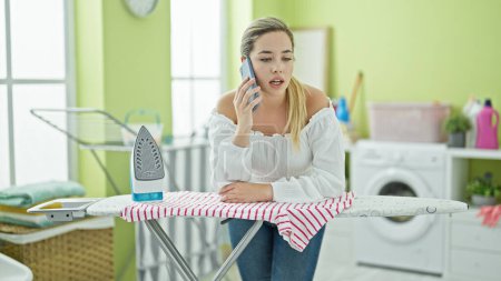 Foto de Mujer rubia joven hablando en el teléfono inteligente apoyado en la tabla de planchar en la sala de lavandería - Imagen libre de derechos