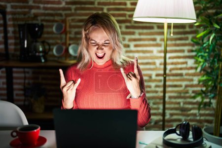 Téléchargez les photos : Femme blonde utilisant un ordinateur portable la nuit à la maison criant avec une expression folle faire symbole rock avec les mains en l'air. star de la musique. concept lourd. - en image libre de droit