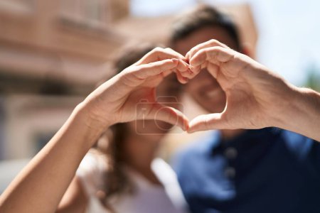 Téléchargez les photos : Jeune couple hispanique souriant confiant faire geste de coeur avec les mains à la rue - en image libre de droit