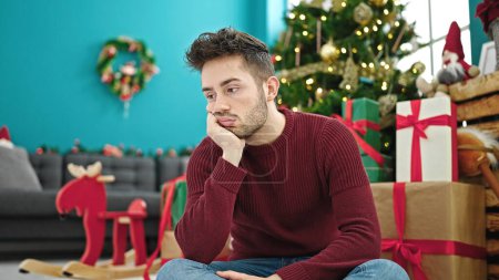 Téléchargez les photos : Jeune homme hispanique célébrant Noël assis sur le sol regardant bouleversé à la maison - en image libre de droit